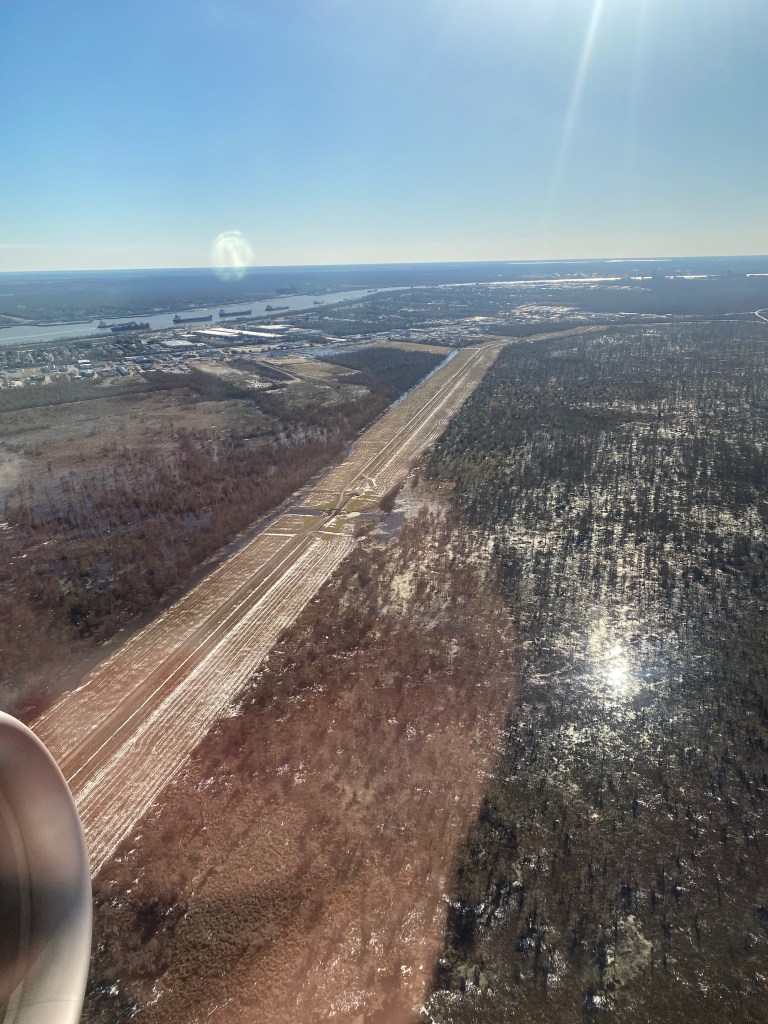 A photograph of Southeast Louisiana taken from a plane window, showing swamps and trees with a road running between them, snow still visible on its surface.