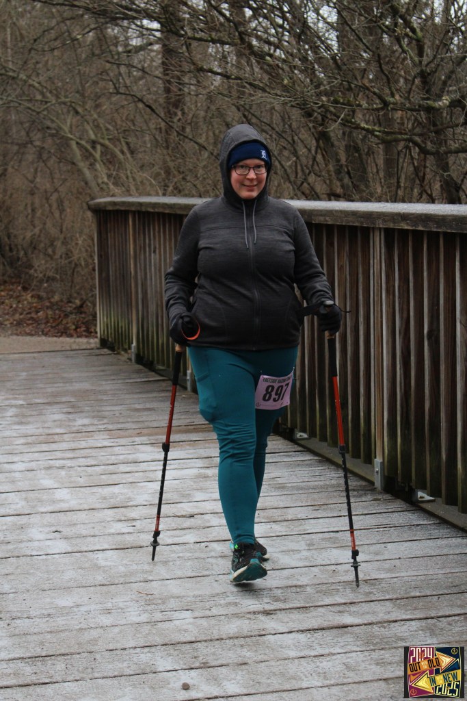 A woman dressed in athletic clothing walks across a wooden bridge while using hiking poles to keep her footing.