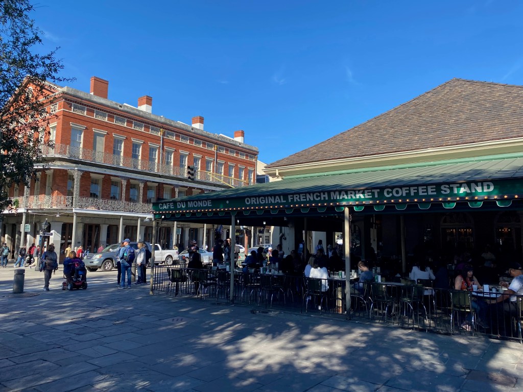 The exterior of the Café du Monde coffee stand in the French Quarter of New Orleans, with crowds of people sitting at tables underneath a green-and-white striped awning.