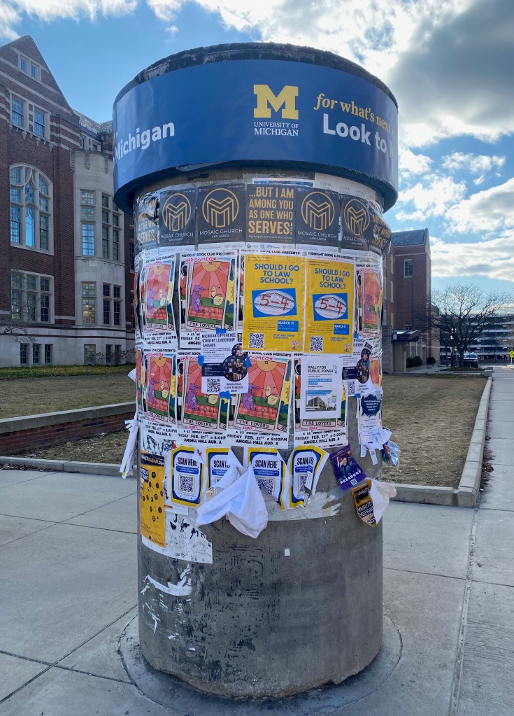 A concrete cylinder at the University of Michigan is covered in flyers and posters, some freshly added, others disintegrating and unreadable.