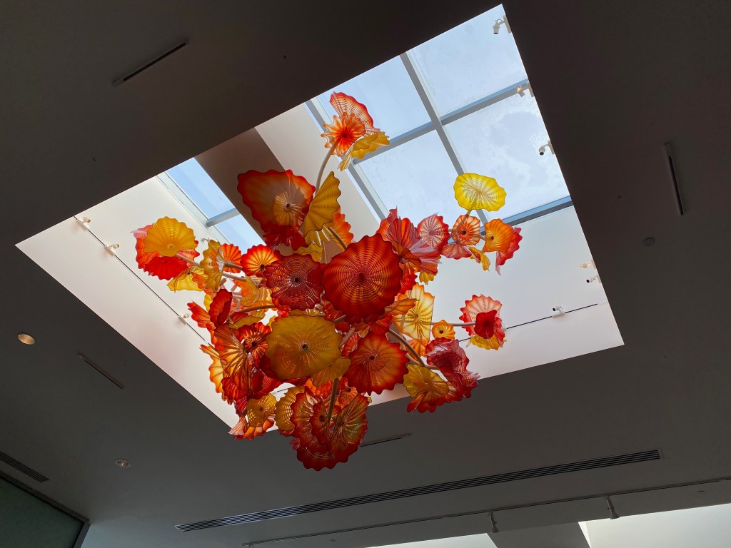 A photograph of a ceiling with a skylight cut into it. A mobile of red, yellow, and orange glass flowers hangs in the skylight well.
