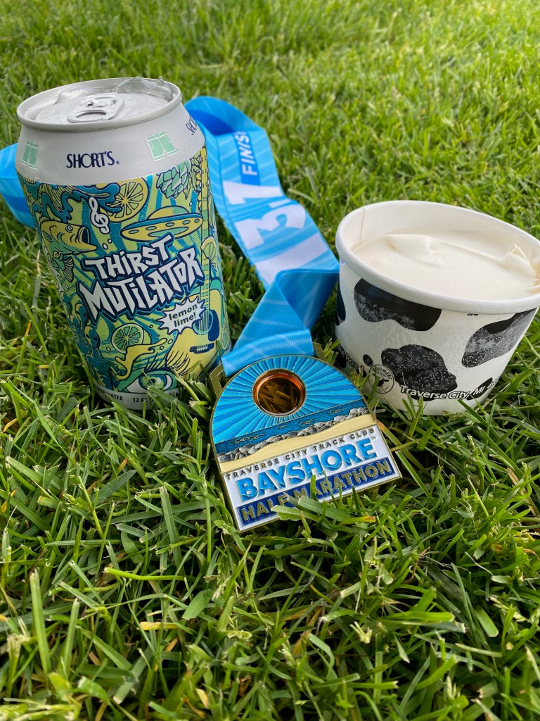 A can of hop water, a medal that reads "Bayshore Half Marathon," and a cup of ice cream sit on a bed of grass.