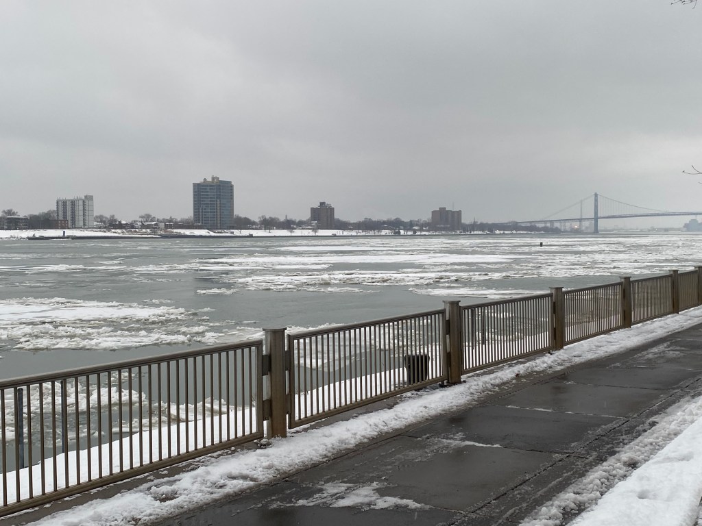 A partially frozen river on a gray, overcast day, with a city skyline and bridge in the background.