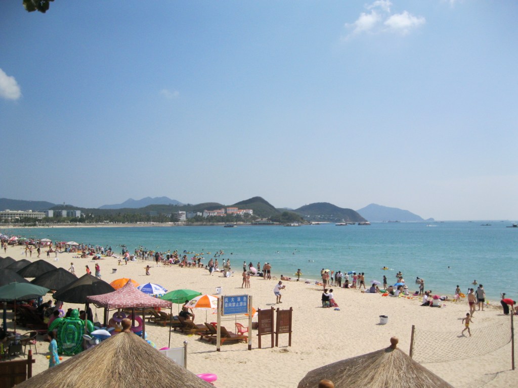 Wide-angle shot of a sunny beach filled with people next to glittering blue-green water.