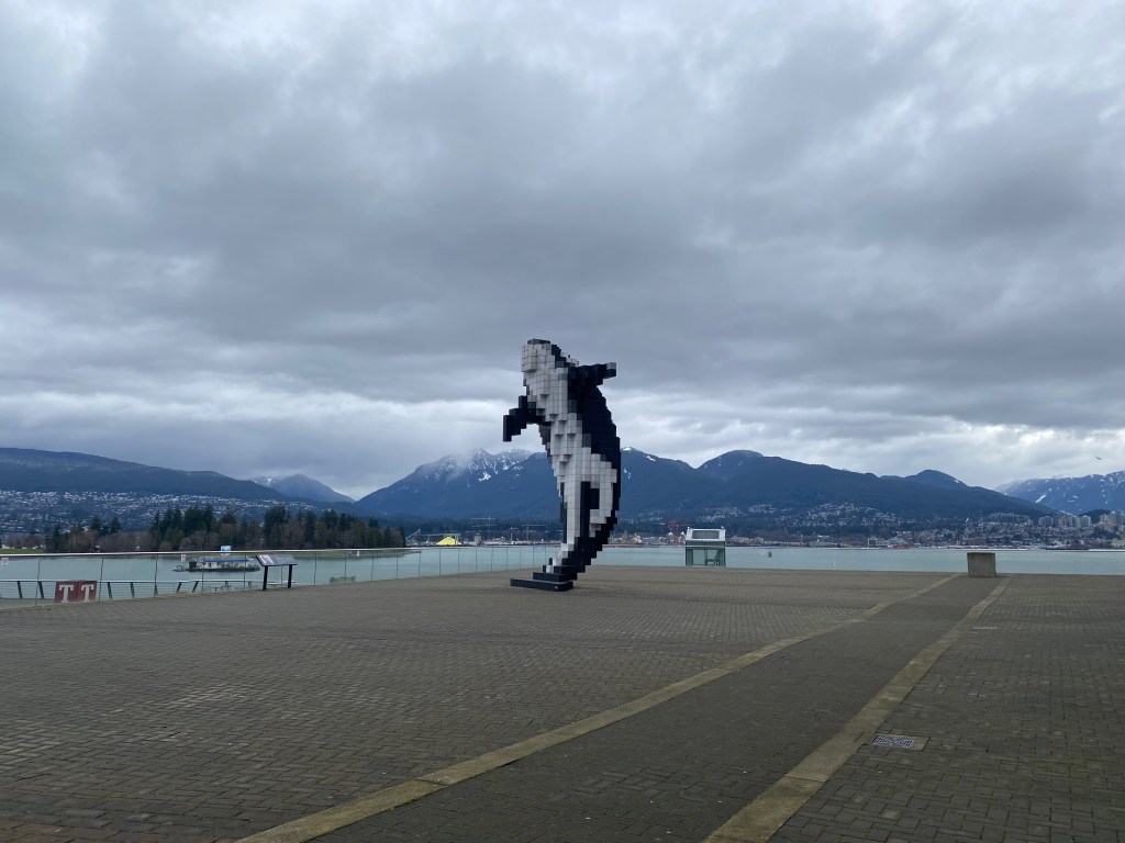 A Lego-style pixelated orca whale sculpture rises from a plaza, with the Vancouver Convention Centre building to its right