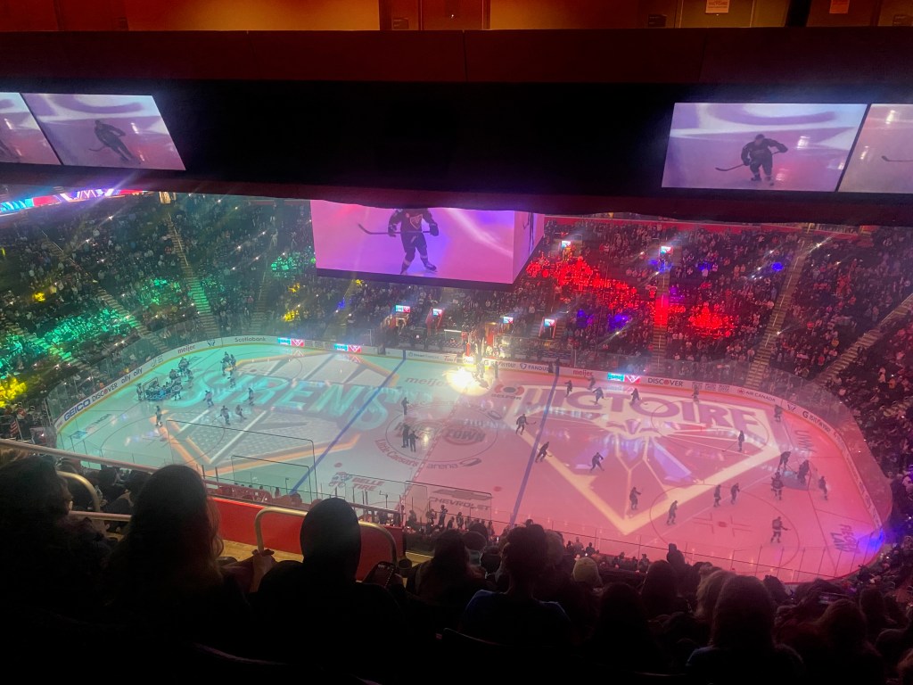 A hockey rink is lit up with projections of logos for the New York Sirens and Montreal Victoire