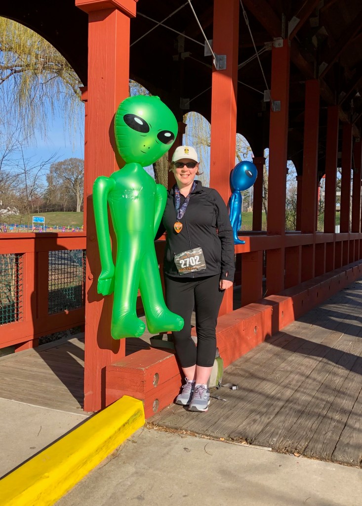 A woman wearing black athletic clothing stands next to an inflatable green alien figure