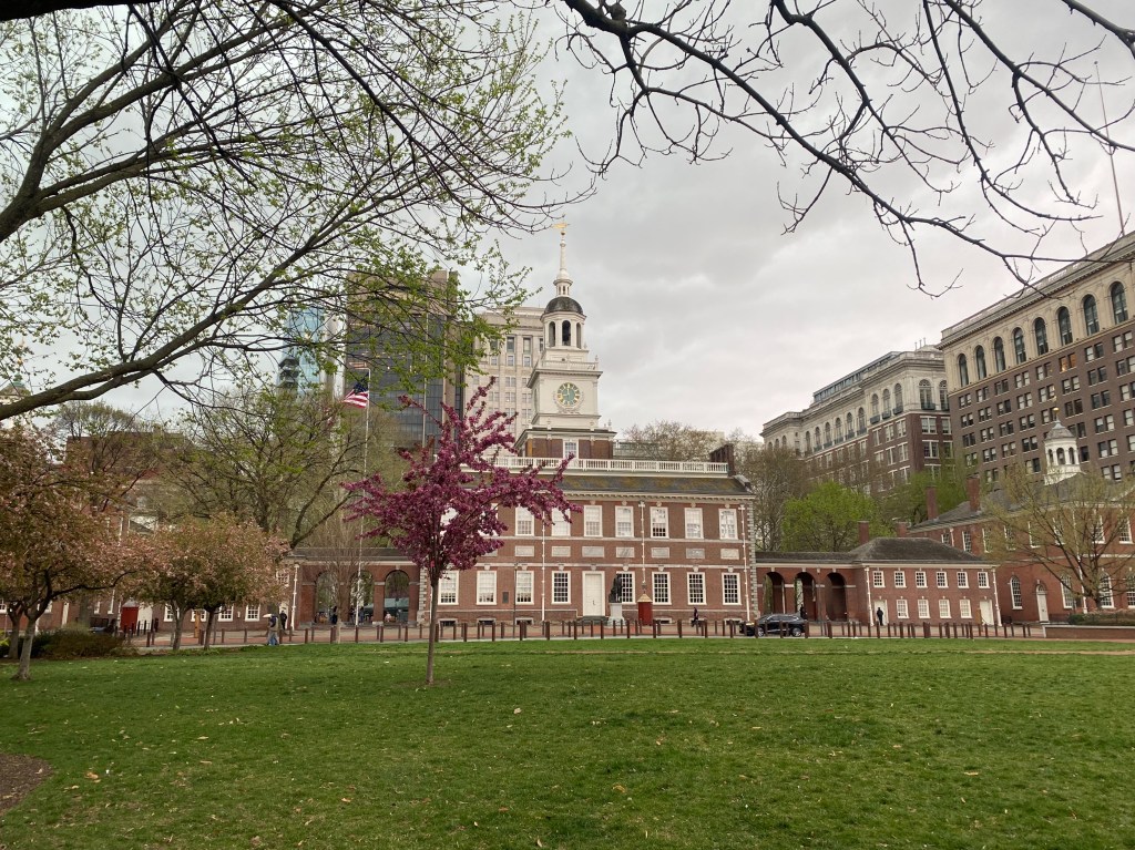 A photograph of Independence Hall, taken from a distance, with a barely flowering tree visible in the foreground.