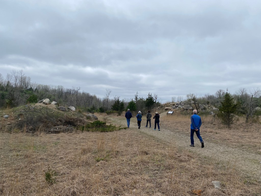 Five people walk along a dirt trail in a large open park, with small grassy hills topped by boulders on both sides of the path.