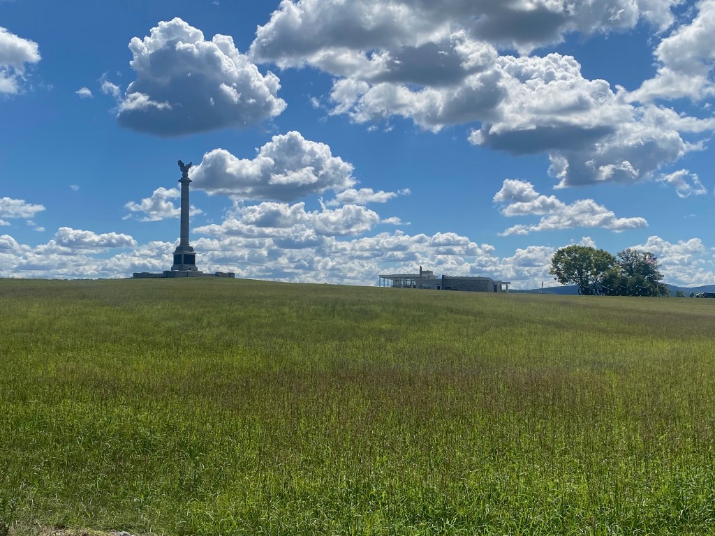A rolling green landscape and blue sky studded with puffy clouds. On the left side of the image is a stone monument.
