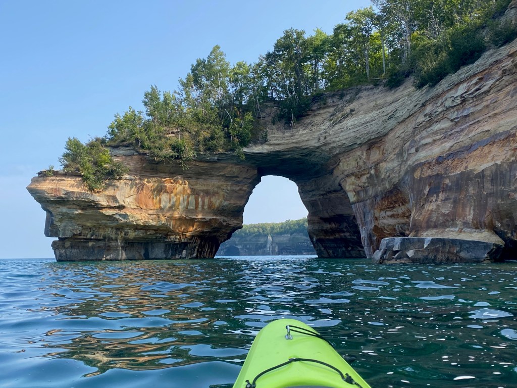 View of a large rock arching over a lake, with the nose of a kayak visible in the foreground.