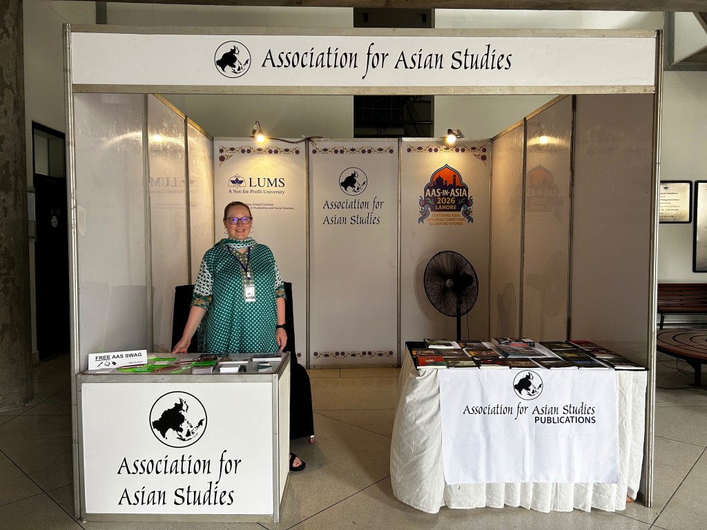 A woman with red hair wearing a green shalwar kameez stands inside an exhibit hall booth labeled "Association for Asian Studies."
