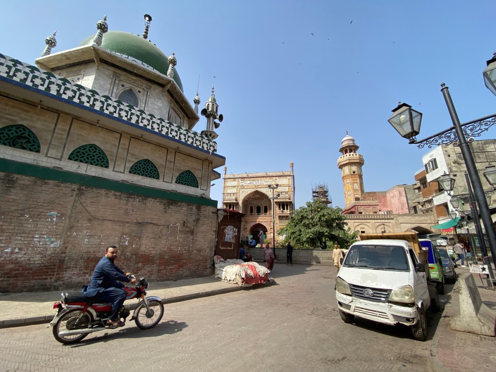 Photograph of a street in Lahore, Pakistan with a mosque in the middle-background and a man on a motorcycle in the foreground.